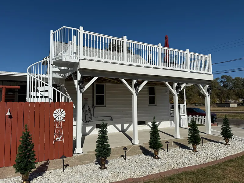 White elevated house with a spacious rooftop deck and ground-level patio with small evergreen trees, Morton, IL Best Construction.
