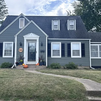 Charming gray house with black shutters, a curved pathway, and autumn decorations.